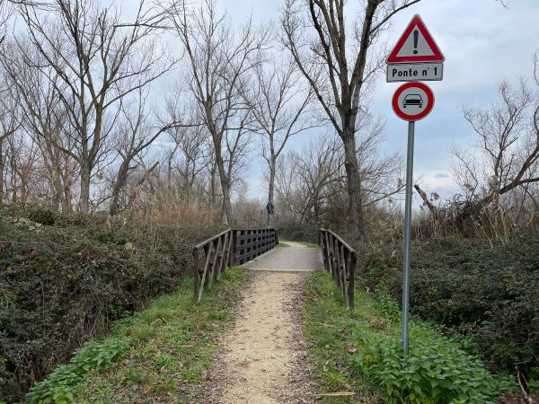 Sentiero sterrato verso ponte in legno con ringhiere di legno. Prima, a destra, segnali stradali con divieto di accesso per auto